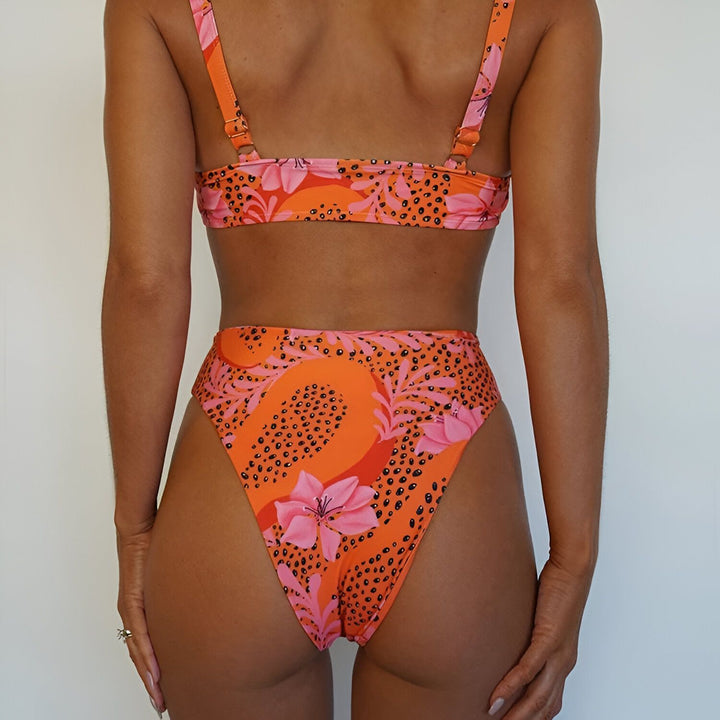 Image d'un ensemble de bikini orange avec des motifs floraux roses et noirs, présenté sur un fond blanc. Le soutien-gorge et la culotte sont visibles, offrant un aperçu du style et du design. Accessoires de plage inclus.
