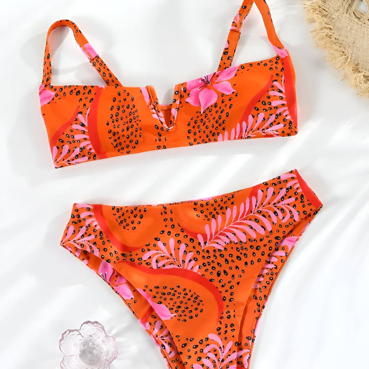 Image d'un bikini orange à motif floral rose et noir, posé à plat sur un fond blanc, avec un chapeau de paille partiellement visible.