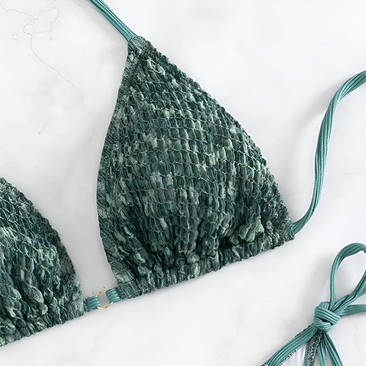 Gros plan d'un bas de bikini vert texturé avec des cordons à nouer sur un fond blanc. Le tissu est plissé et ressemble à un motif de peau de serpent. Une partie du haut de bikini est visible en arrière-plan.