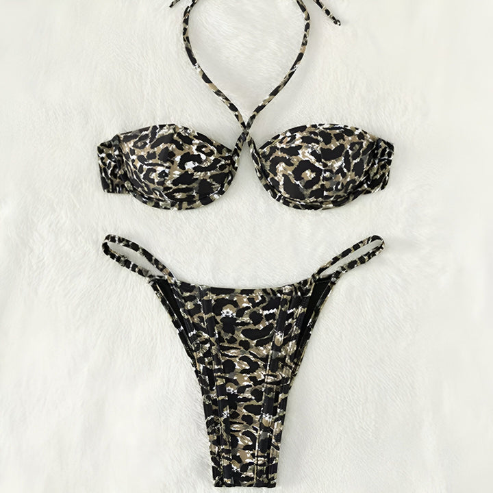 Une femme blonde porte un bikini léopard. Haut bandeau avec bretelles croisées, bas brésilien. Bijou au nombril et bracelet doré. Fond blanc uni.