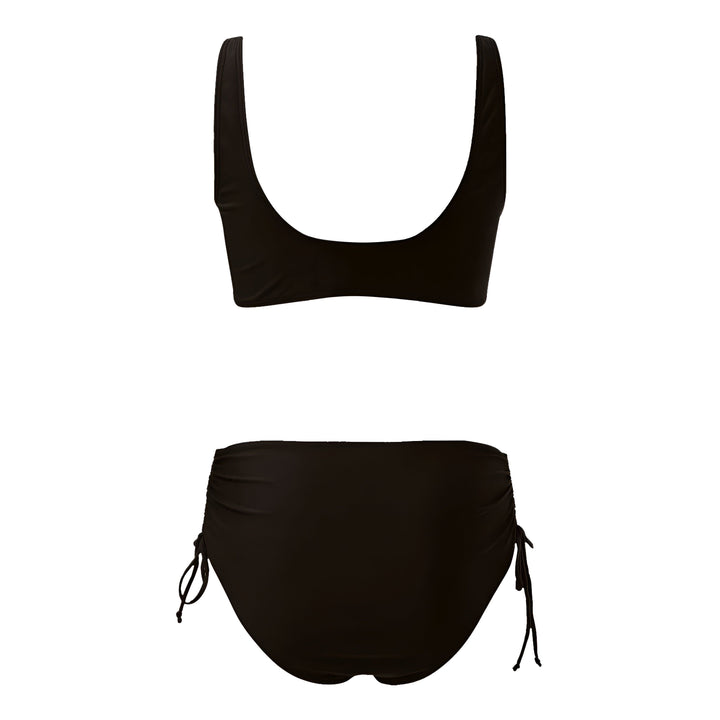 Une femme en maillot de bain noir deux pièces avec un haut croisé et un bas taille haute à nouer, posant devant l'océan avec un collier blanc.