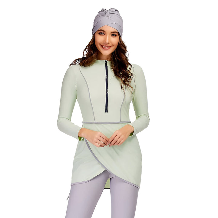 Une femme souriante porte un maillot de bain burkini vert d'eau et gris avec un turban assorti, sur un fond blanc uni.