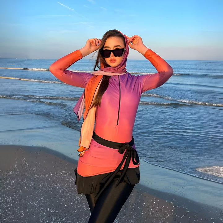 Femme portant un burkini rose et orange avec un foulard assorti et des lunettes de soleil noires sur une plage ensoleillée.