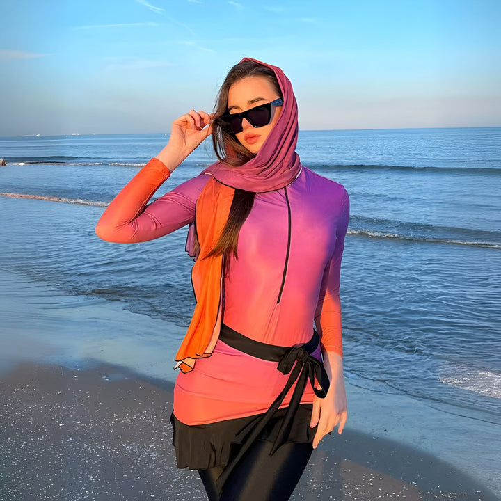 Femme voilée en maillot de bain intégral coloré sur une plage ensoleillée. Elle porte des lunettes de soleil et pose avec confiance, la mer à l'arrière-plan. Mode de bain pudique et élégante.