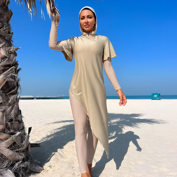 Femme portant un burkini beige avec capuche sur une plage de sable blanc, dos à la caméra. Un palmier est visible sur la gauche, avec un ciel bleu clair et la mer en arrière-plan. Mode de plage modeste.