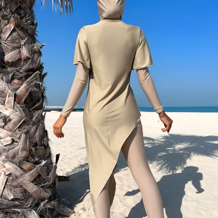 Femme sur la plage portant un maillot de bain burkini beige, foulard de tête beige assorti, avec un palmier sur le côté gauche, ciel bleu clair et mer à l'arrière-plan.