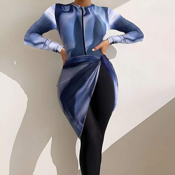 Femme portant un maillot de bain bleu et noir avec une fermeture éclair et une jupe nouée, devant un fond blanc. Mode de plage tendance et confortable.