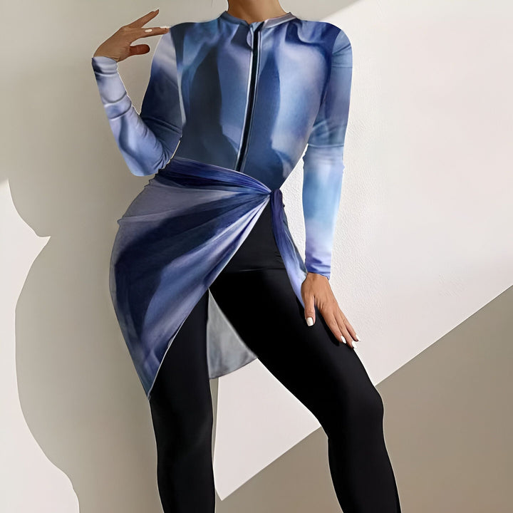 Femme portant un justaucorps imprimé bleu et blanc, leggings noirs et paréo assorti. Ses mains sont posées sur ses hanches. Lumière naturelle douce sur fond blanc.