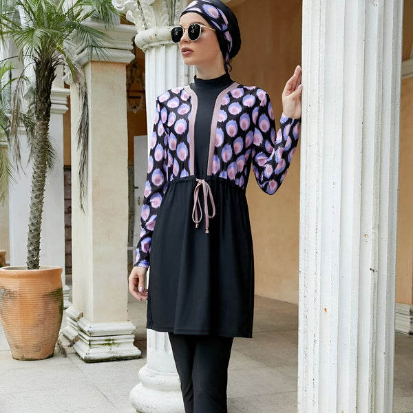 Femme portant un maillot de bain burkini noir avec une veste à motifs violets et roses, debout à côté de colonnes blanches et de palmiers. Vêtements de bain islamiques, mode pudique.
