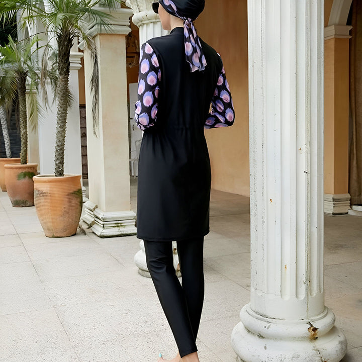 Femme portant un maillot de bain burkini noir avec manches longues à motif floral violet, avec un hijab assorti, posant devant des colonnes blanches et des palmiers.