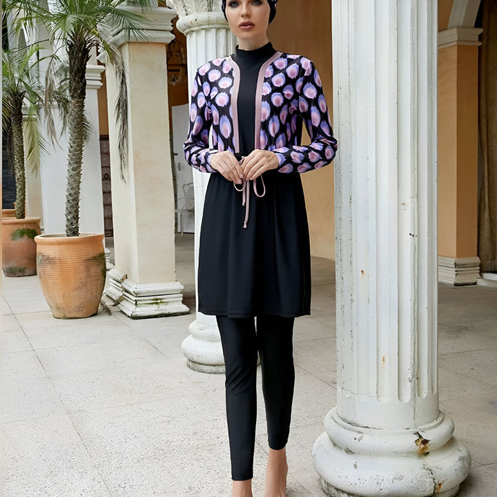 Femme portant un maillot de bain burkini noir avec un imprimé coloré, des lunettes de soleil et un hijab assorti, adossée à une colonne blanche dans un cadre extérieur ensoleillé.