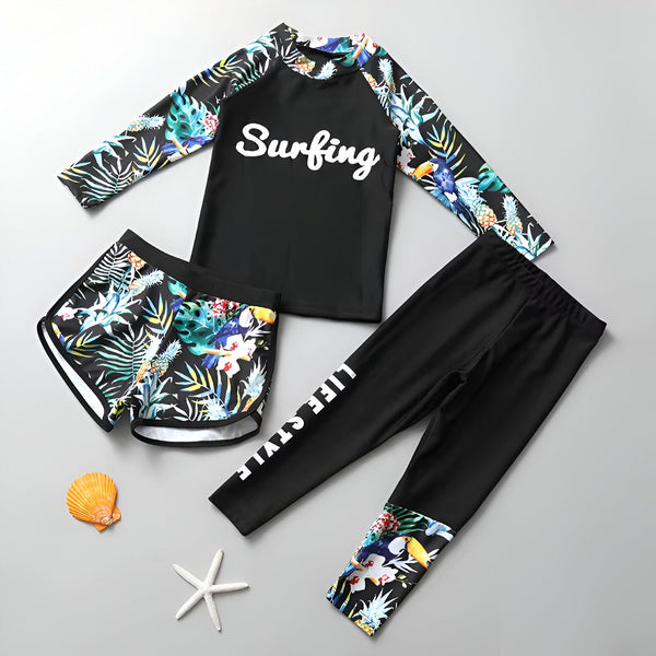 Maillot de bain anti-UV noir à manches longues avec motif tropical (feuilles, ananas, toucan). Le mot "Surfing" est écrit en blanc sur le devant.