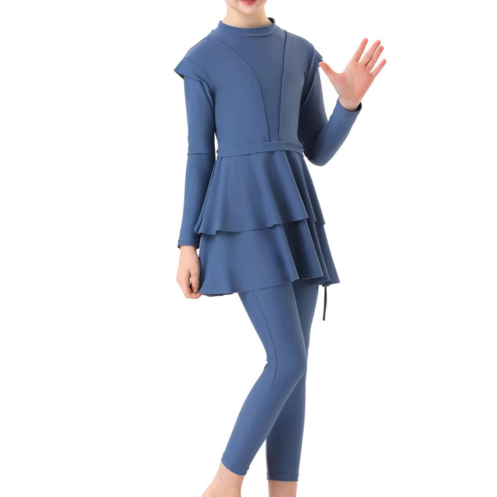 Fille portant un maillot de bain burkini bleu à manches longues, deux pièces, avec une jupe à volants. Mode de bain pudique.