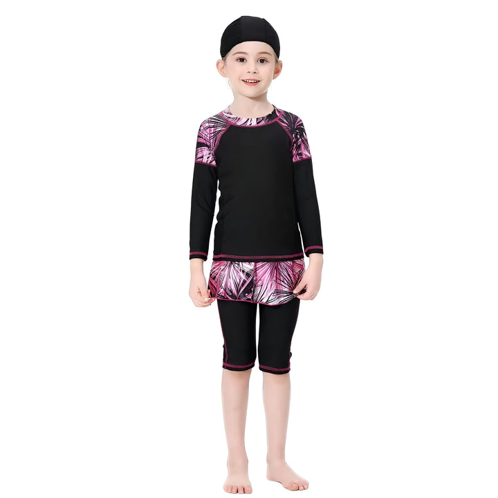Une fillette souriante porte un maillot de bain modeste noir et rose avec des motifs tropicaux. Il comprend un haut à manches longues et un bas de type legging avec une jupe intégrée.