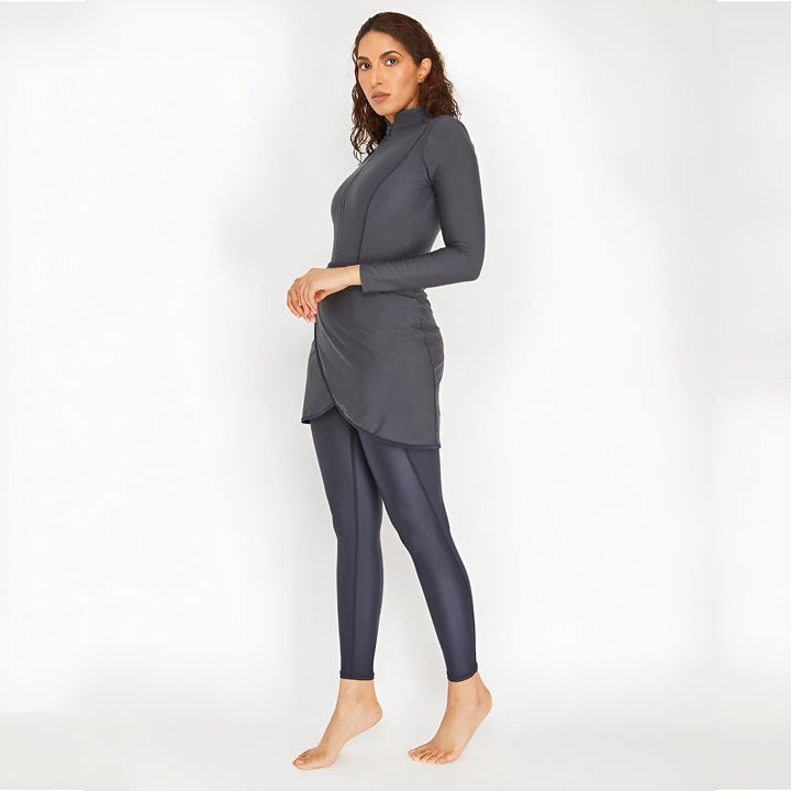 Femme portant un maillot de bain burkini gris avec un haut à manches longues zippé et une jupe portefeuille, sur un fond blanc uni.