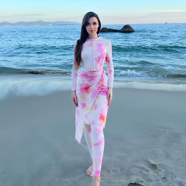 Femme de dos, vêtue d'une robe longue tie-dye rose et blanche, sur une plage avec vue sur l'océan et les montagnes au loin.