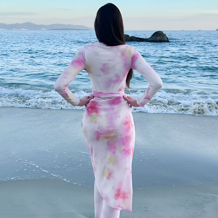 Femme sur la plage en tenue tie-dye rose et blanche. Mer et montagne en arrière-plan. Mode balnéaire, style estival, vacances à la mer.