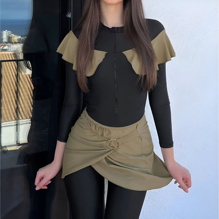 Femme brune posant sur un escalier blanc, portant un maillot de bain couvrant noir avec des détails kaki. Style moderne et élégant pour la plage.