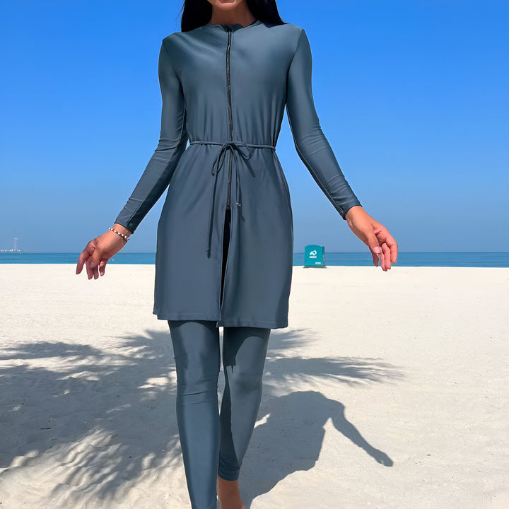 Femme en tenue de sport bleu-gris sur plage de sable blanc avec ciel bleu et mer turquoise. Look fitness, bien-être et vacances.