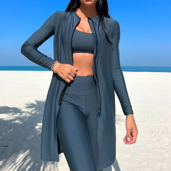 Femme sur la plage, portant un maillot de bain de protection gris avec fermeture éclair, un haut et un legging assortis. Bracelet fleuri au poignet. Ciel bleu et mer turquoise en arrière-plan. Mode de bain pudique.