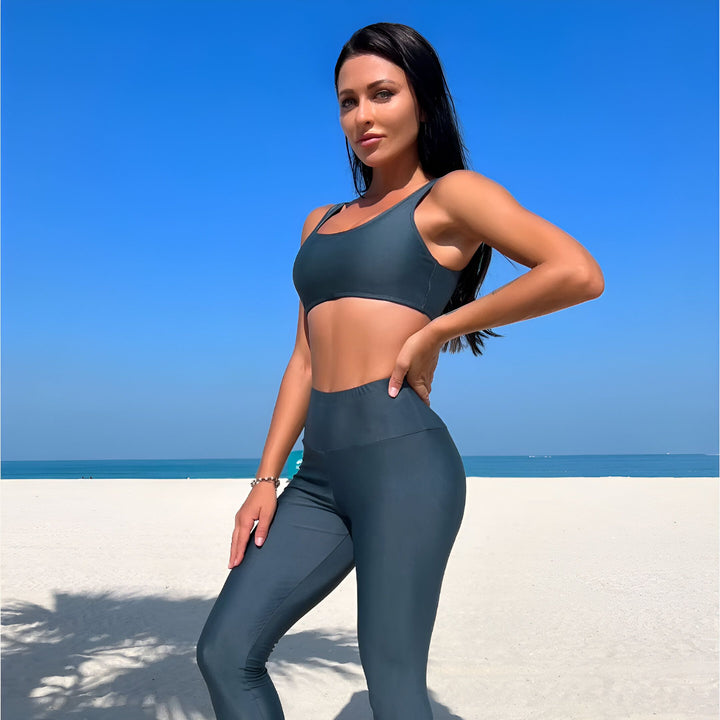 Femme portant un burkini gris sur une plage de sable blanc, ciel bleu en arrière-plan. Vêtement de bain couvrant avec fermeture éclair et ceinture. Mode pudique.