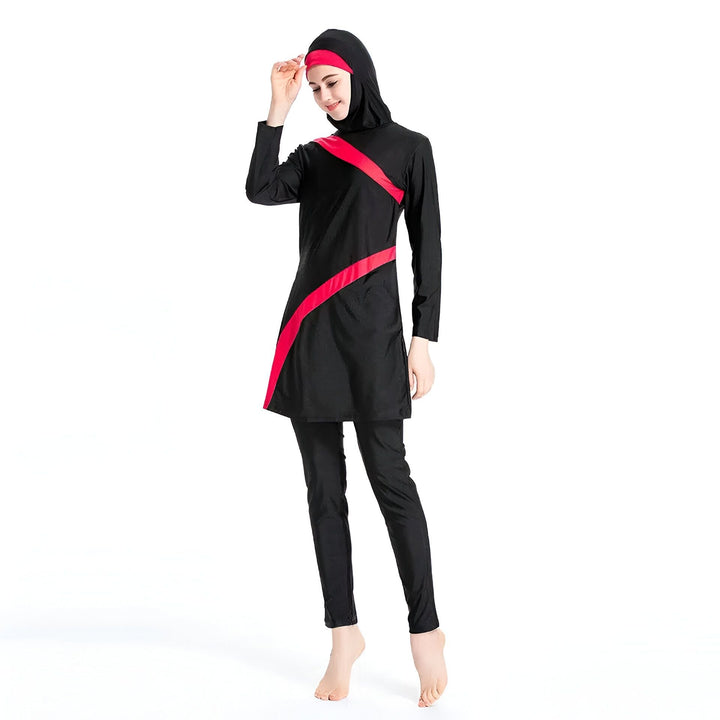 Une femme porte un burkini noir avec une bande verte diagonale. Le burkini comprend un haut à manches longues, un pantalon et un hijab assorti. Elle est pieds nus sur un fond blanc.