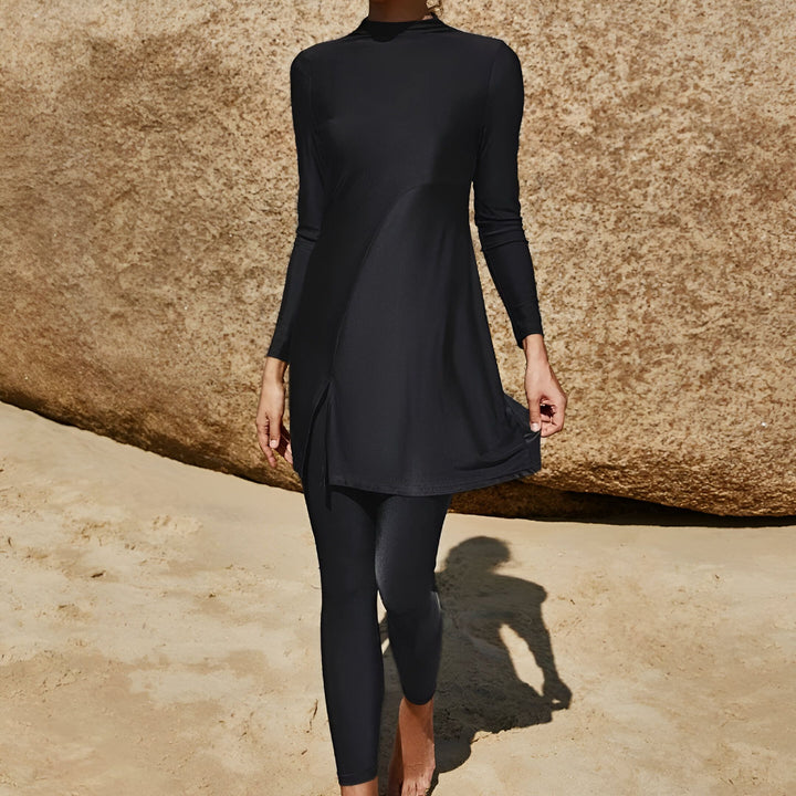 Femme portant un maillot de bain burkini noir, posant devant des rochers sur la plage. Elle porte des lunettes de soleil dorées.