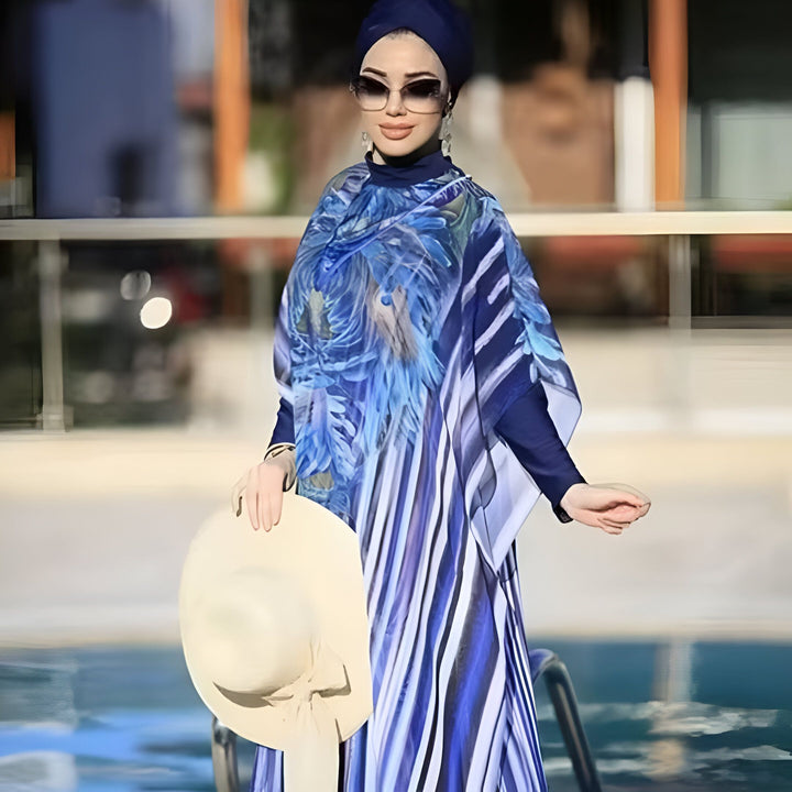 Une femme souriante portant un burkini bleu floral, des lunettes de soleil, un turban assorti et tenant un chapeau de soleil, debout près d'une piscine.