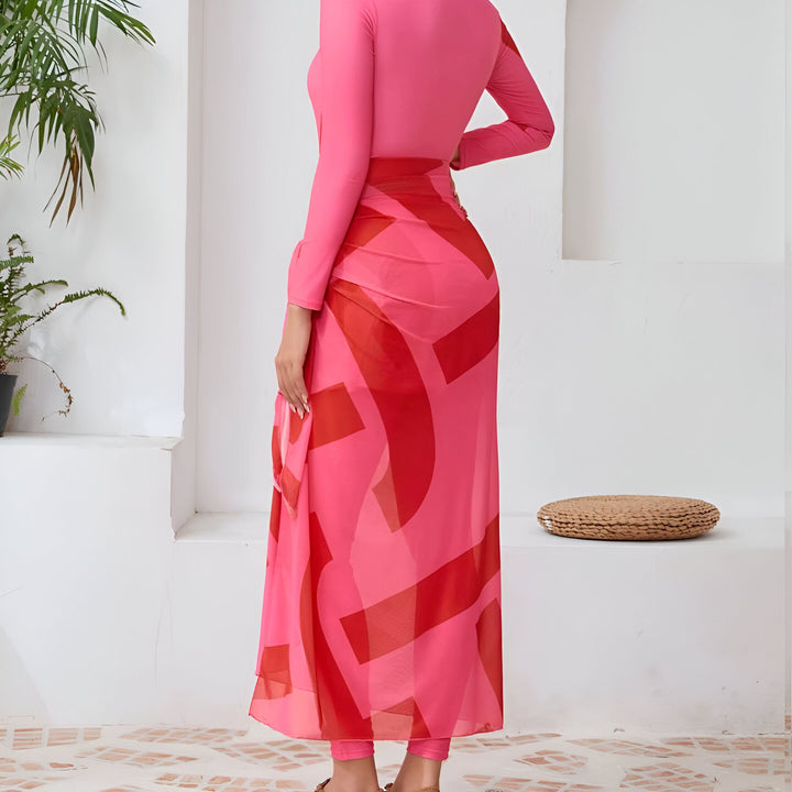 Femme souriante portant un maillot de bain rose à manches longues et un paréo à motifs géométriques, chapeau de paille à larges bords, posant dans un intérieur minimaliste avec des plantes. Mode estivale, style de plage.