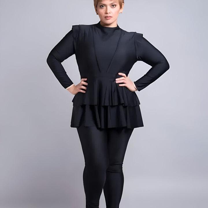 Femme portant un maillot de bain intégral noir avec un haut à volants et un pantalon de bain moulant, mains sur les hanches, fond gris uni.