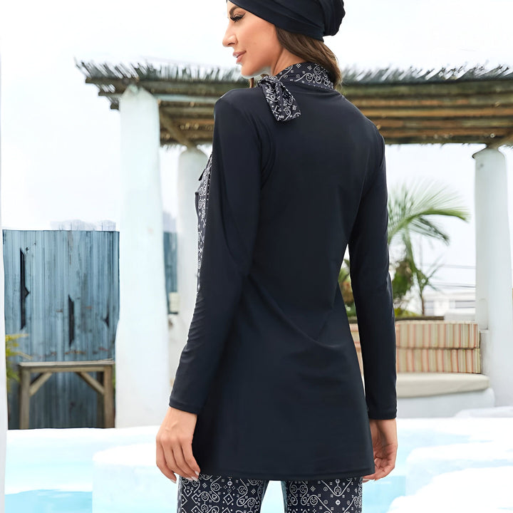Femme portant un maillot de bain pudique noir avec un foulard et un bonnet assortis, vue de dos, près d'une piscine. Le maillot a des détails imprimés en blanc et noir.