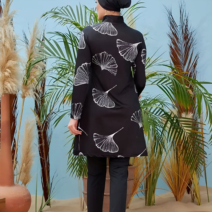 Maillot de bain intégral noir avec motifs de feuilles blanches, vu de dos. Porté avec un bonnet de bain noir. Arrière-plan avec des plantes tropicales et un mur bleu. Vêtements de bain modestes.