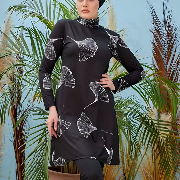 Une femme portant un maillot de bain intégral noir à motifs de feuilles blanches et un hijab assorti, posant devant des palmiers. Mode pudique, vêtements de bain islamiques.