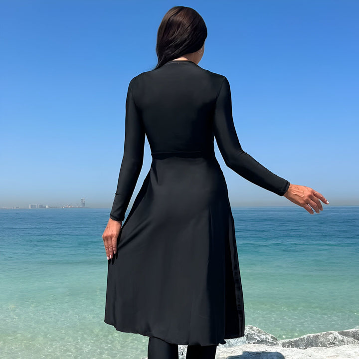 Vue arrière d'une femme portant un maillot de bain intégral noir (burkini) sur une plage ensoleillée avec un ciel bleu et l'océan turquoise. Mode de bain modeste, protection solaire, vêtements de plage islamiques.