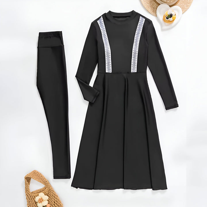 Une femme élégante en maillot de bain modeste noir avec des détails bleus et blancs, posant sur des rochers près de la mer turquoise. L'arrière-plan montre un ciel bleu clair et des bâtiments lointains.