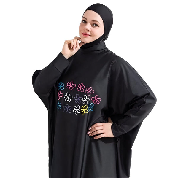 Femme portant un burkini noir à manches longues avec motifs floraux colorés et un hijab, posant devant un fond blanc avec une plante verte. Vêtement de bain modeste.