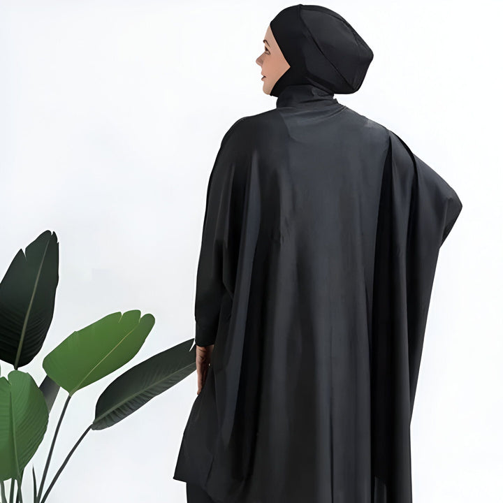 Une femme vue de dos porte un burkini noir avec un hijab assorti, posant à côté d'une plante verte sur fond blanc. Mode de bain pudique.