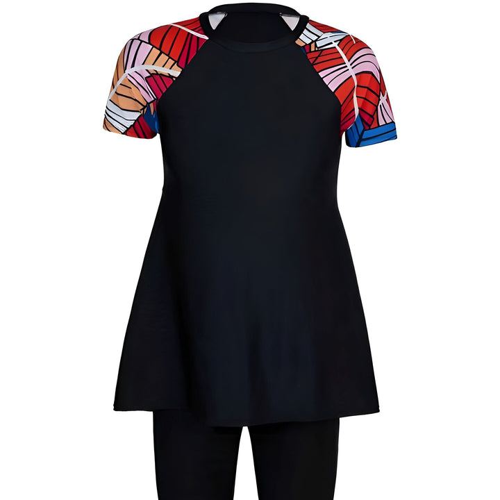 Femme portant un maillot de bain noir avec des manches à motifs floraux colorés, posant devant un ciel bleu nuageux sous un chapeau de paille et des feuilles de palmier.