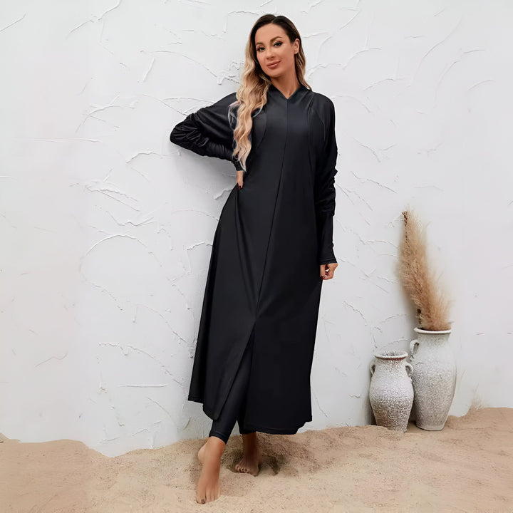 Une femme portant un maillot de bain intégral noir élégant, posant devant un mur blanc texturé. Des vases décoratifs et du sable ajoutent une touche estivale à la scène. Vêtements de bain modestes pour femmes.