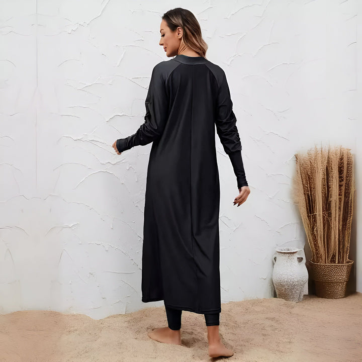 Femme portant une burkini noire, un maillot de bain intégral pour femmes musulmanes. Elle pose devant un mur texturé blanc et des vases décoratifs. Vêtements de bain islamiques, mode modeste, burkini élégante.