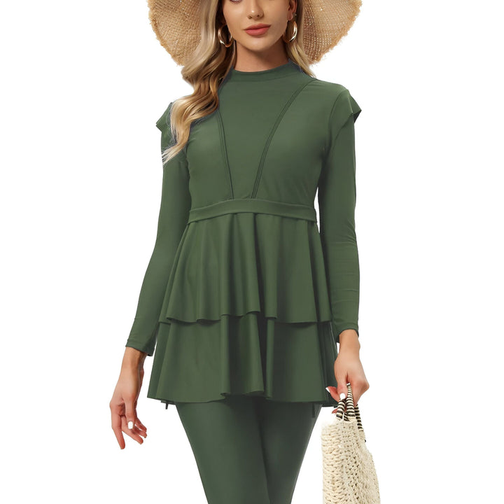 Femme portant un maillot de bain modeste vert olive avec haut à volants, manches longues et chapeau de paille, tenant un sac de plage. Mode plage pudique et élégante.