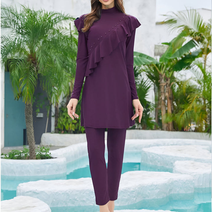 Femme portant un maillot de bain intégral prune avec des volants et des perles, devant un mur blanc avec une fenêtre bleue et des plantes. Vêtement de bain modeste et élégant pour femmes musulmanes.