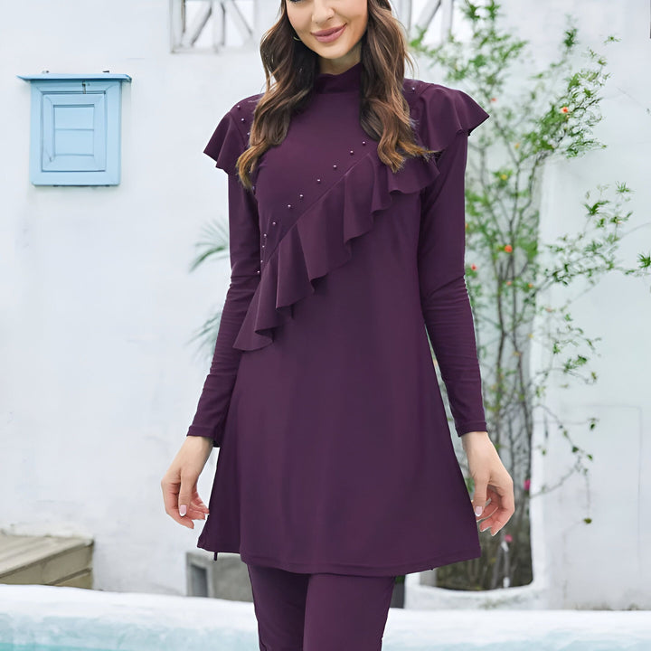 Une femme souriante porte un maillot de bain modeste violet avec des détails à volants et des perles. Elle se tient devant un mur blanc avec une petite fenêtre bleue.