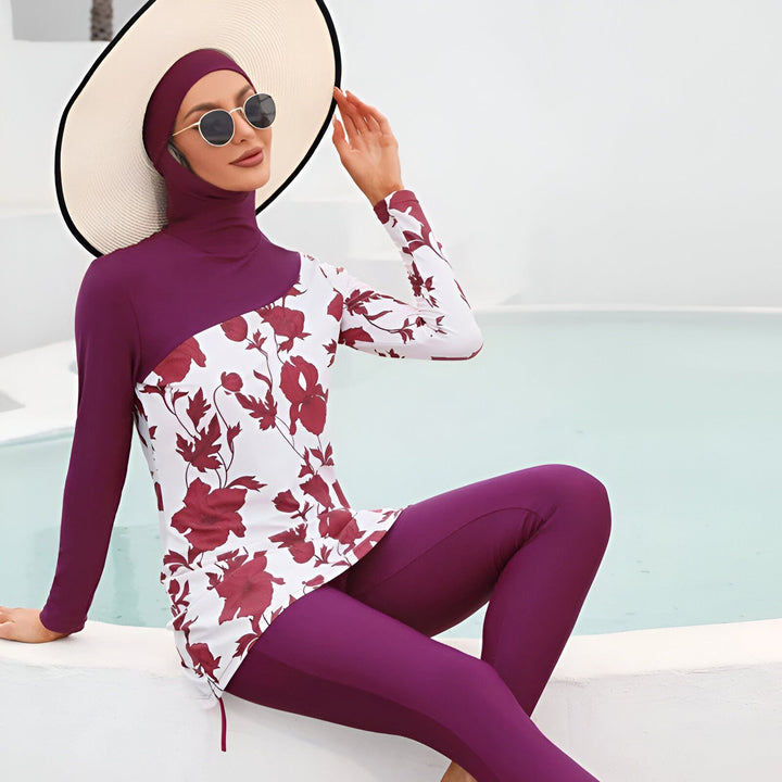 Femme portant un burkini violet et blanc à motif floral, lunettes de soleil rondes, chapeau de paille à bord large, assise au bord d'une piscine. Mode de bain pudique, style et protection solaire.