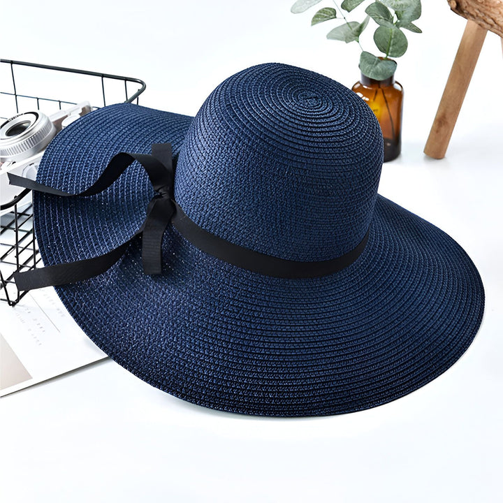Chapeau de soleil bleu marine avec un ruban noir, sur une table avec une décoration florale discrète. Accessoire de mode estivale élégant et pratique.