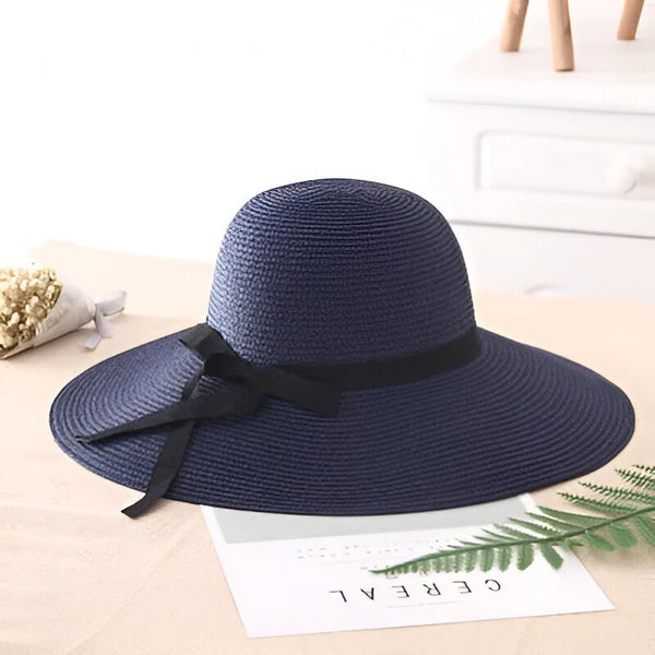Chapeau de soleil bleu marine en paille avec ruban noir, posé sur une surface blanche avec des accessoires en arrière-plan. Accessoire de mode pour l'été, protection solaire élégante.