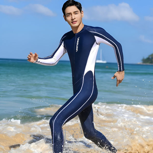 Un homme portant une combinaison de plongée bleue et blanche, debout sur une plage ensoleillée, avec l'océan en arrière-plan. Il porte des lunettes de soleil vertes.