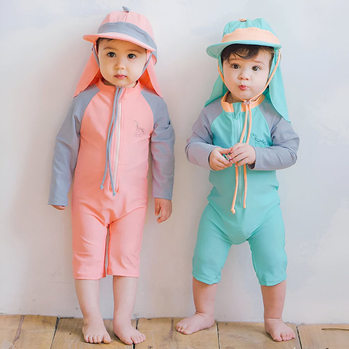 Portrait d'un bébé portant un chapeau de soleil rose et bleu avec protection de nuque. Le chapeau a des détails en forme d'écailles de dinosaure sur le dessus.