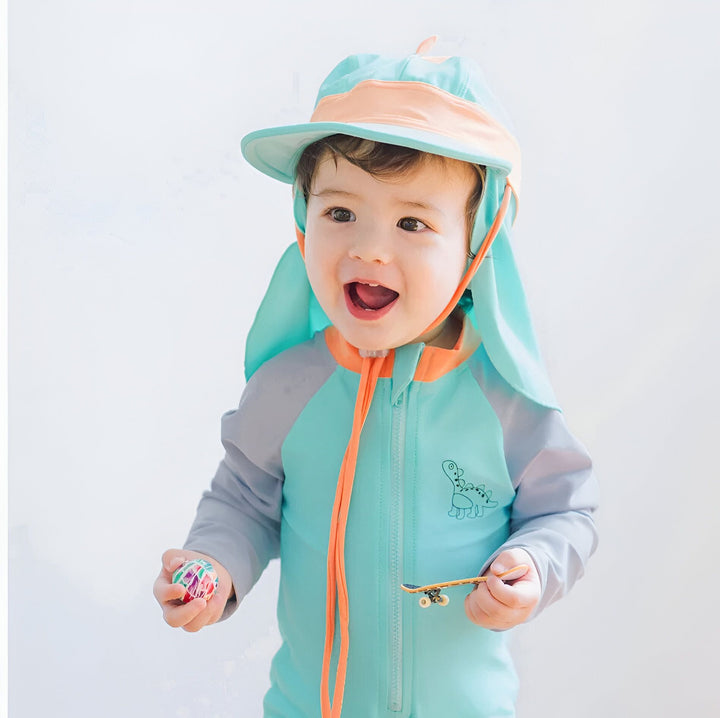 Un bébé portant un maillot de bain une pièce rose et gris avec un chapeau assorti. Il regarde vers le haut avec une expression curieuse. Protection solaire pour enfant.