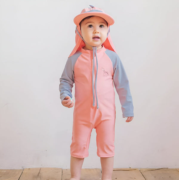 Un bébé souriant, portant un maillot de bain turquoise à manches grises avec un dessin de dinosaure, un chapeau de soleil assorti et tenant une balle et un mini skateboard.